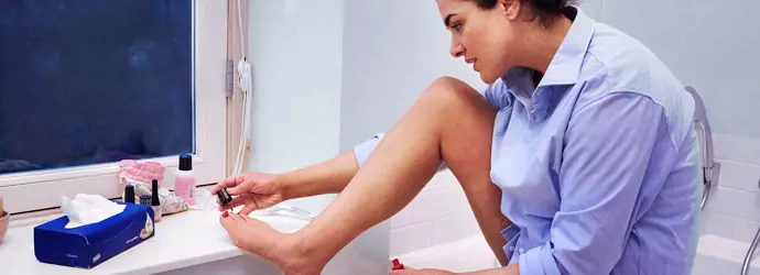 Een vrouw lakt haar teennagels in een badkamer; een doos tissues ligt voor haar.
